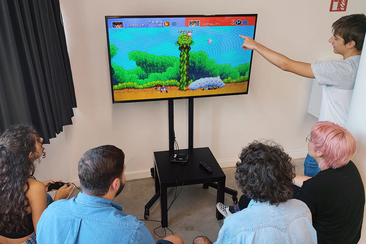 A group of 5 students play a Sega Mega Drive game around a tv.