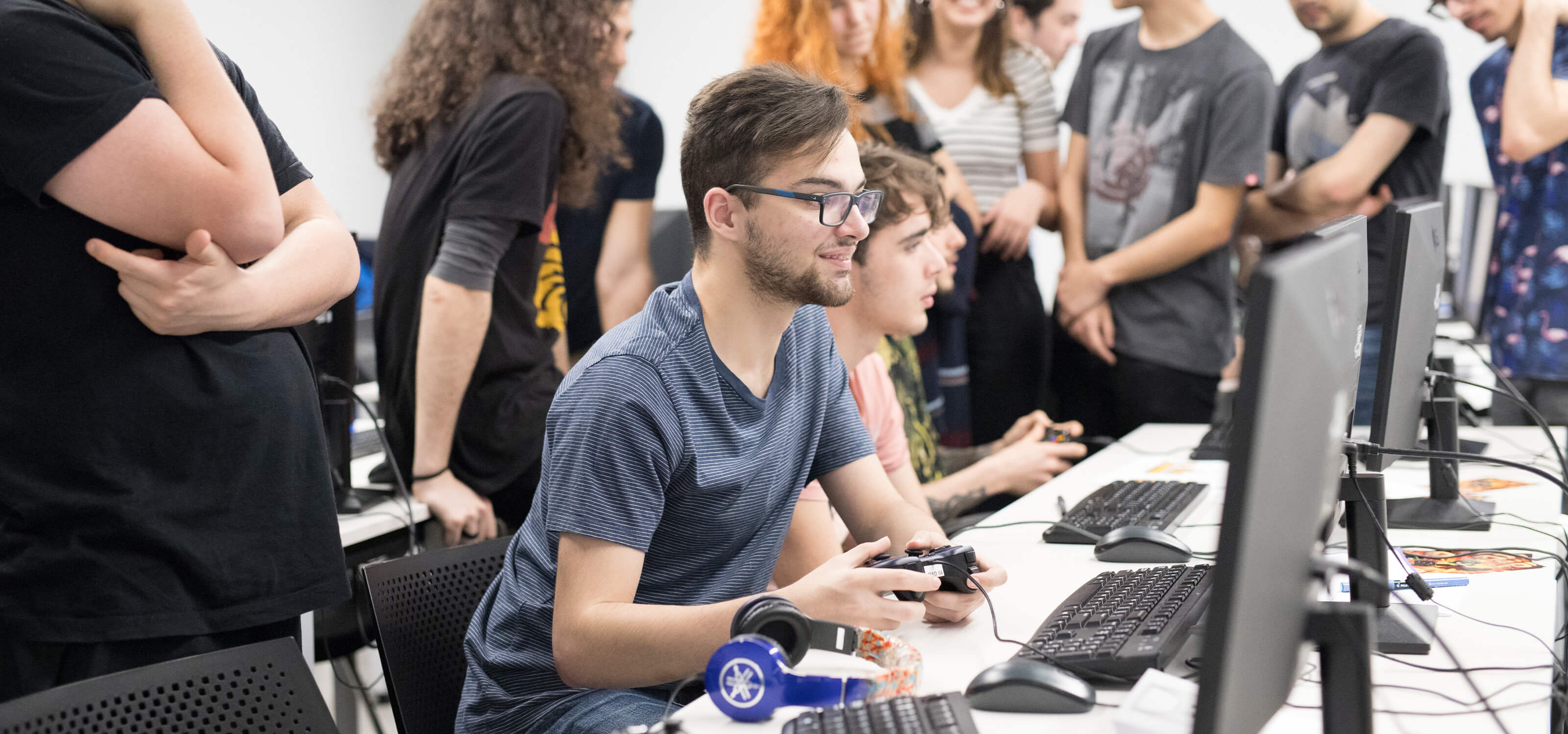 A student holds a controller and watches the monitor in front of him as his classmates surround the table.