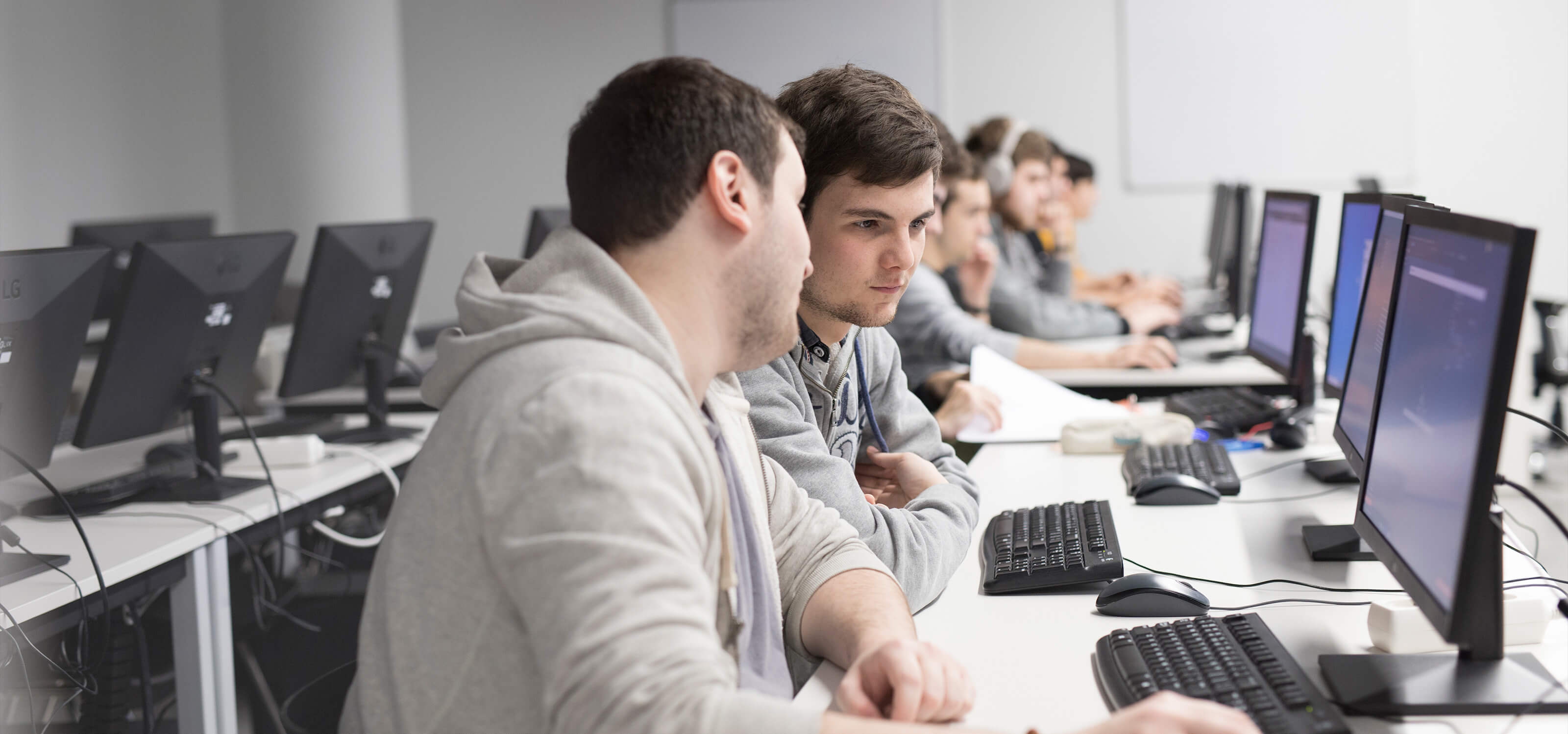 Dos estudiantes trabajan juntos mientras están sentados en una mesa llena de ordenadores con otros compañeros de clase.