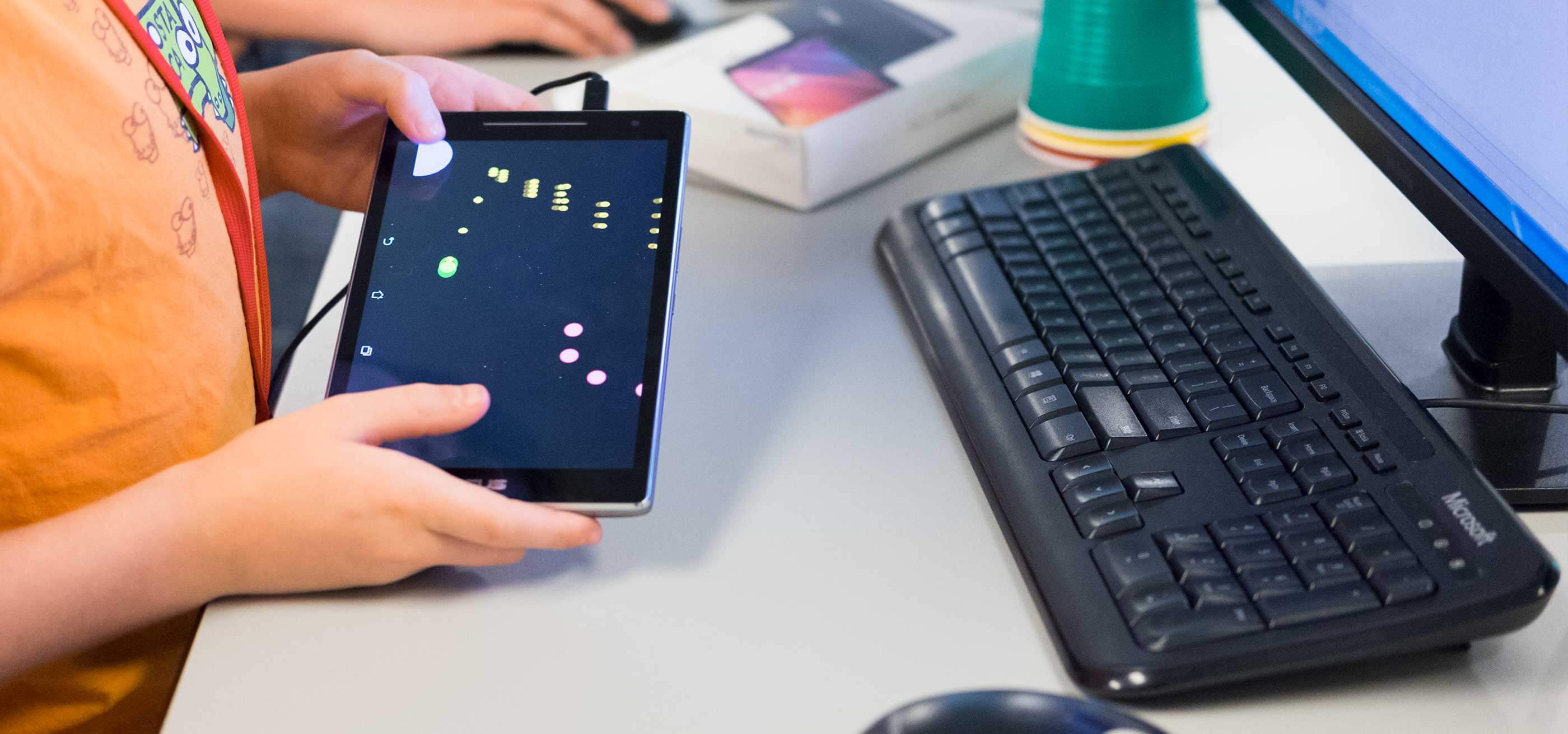 Un estudiante sostiene una tableta con un juego móvil junto a un teclado de ordenador