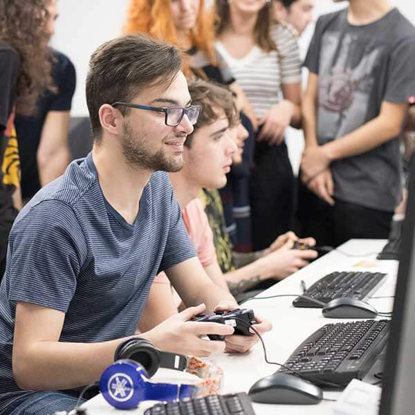 A student holds a controller and watches the monitor in front of him as his classmates surround the table