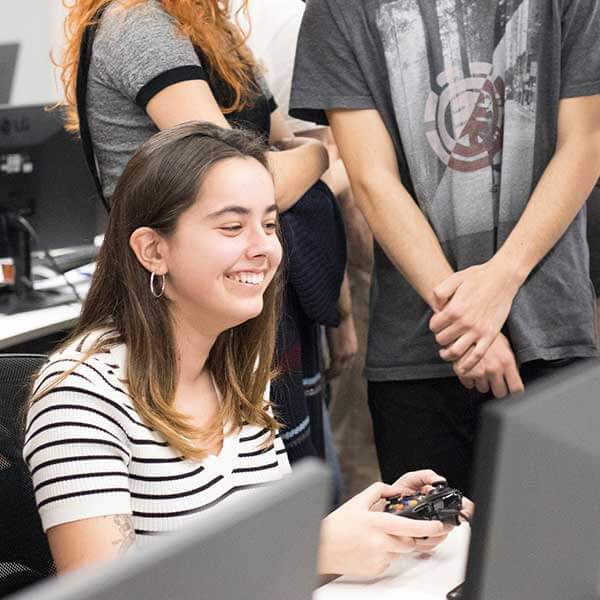 Los estudiantes se reúnen alrededor de un monitor para ver a una compañera de clase jugar a su juego