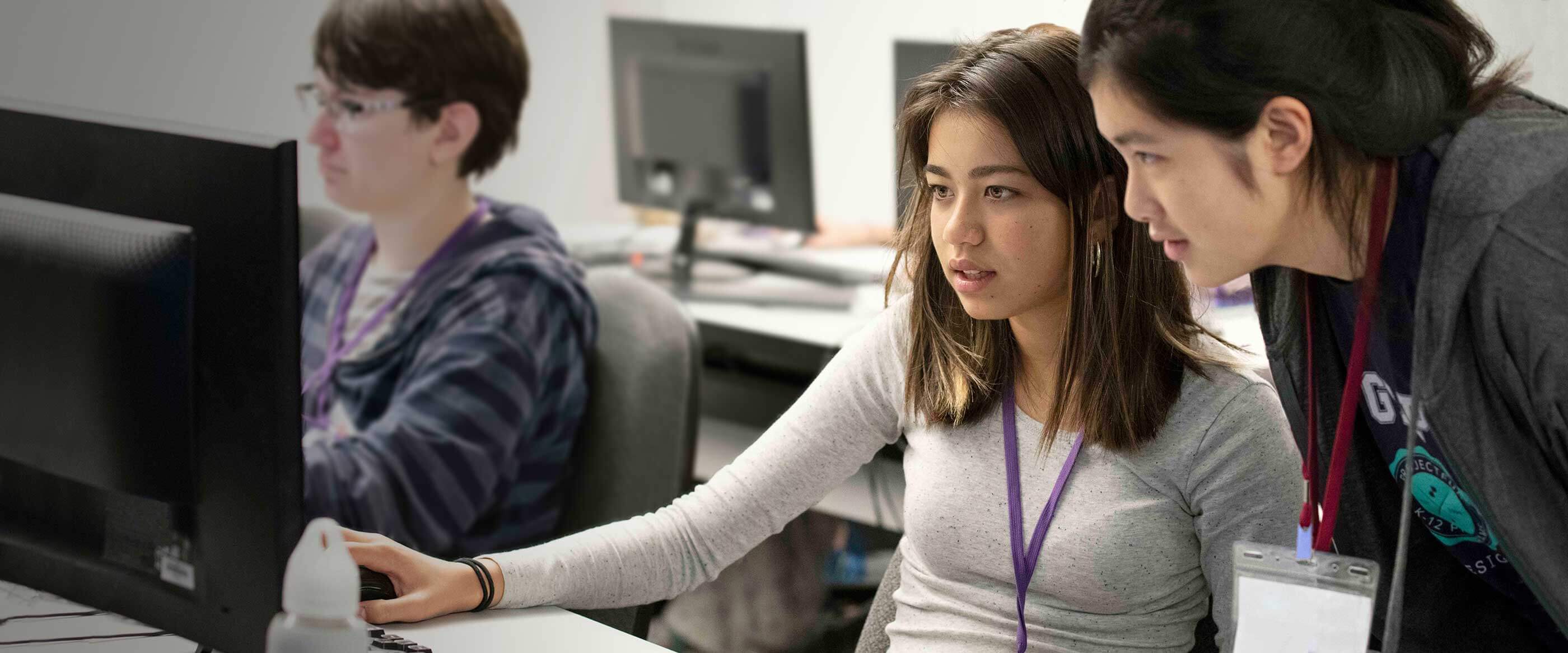 Una alumna sentada frente a un monitor de ordenador con un instructor ayudandole mientras se apoya en su hombro