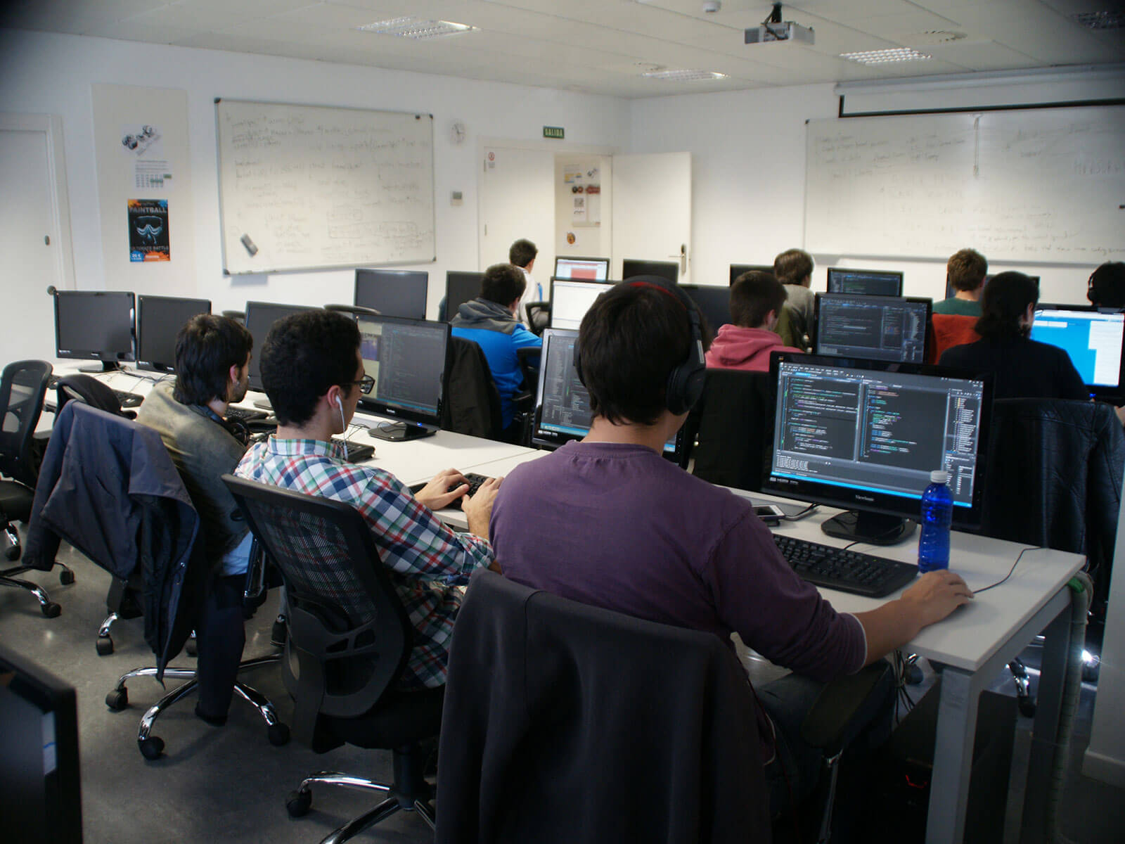 Multiple students sit in at computer workstations while working on programming