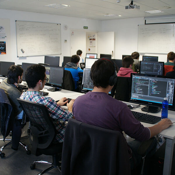 Multiple students sit in at computer workstations while working on programming