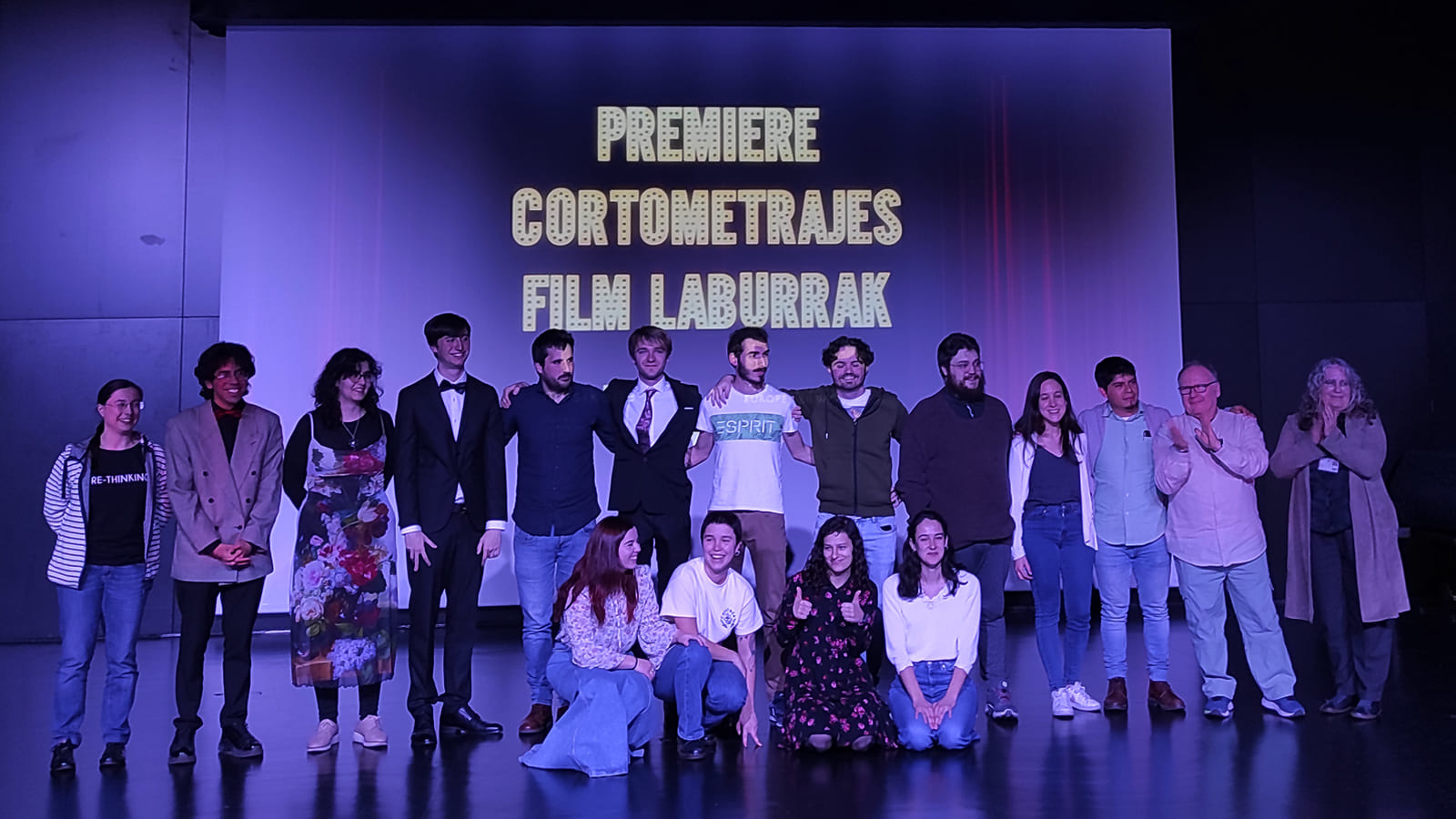  A group of DigiPen and Musikene students standing and posing for a photograph in front of a projection screen showcasing a film premiere.