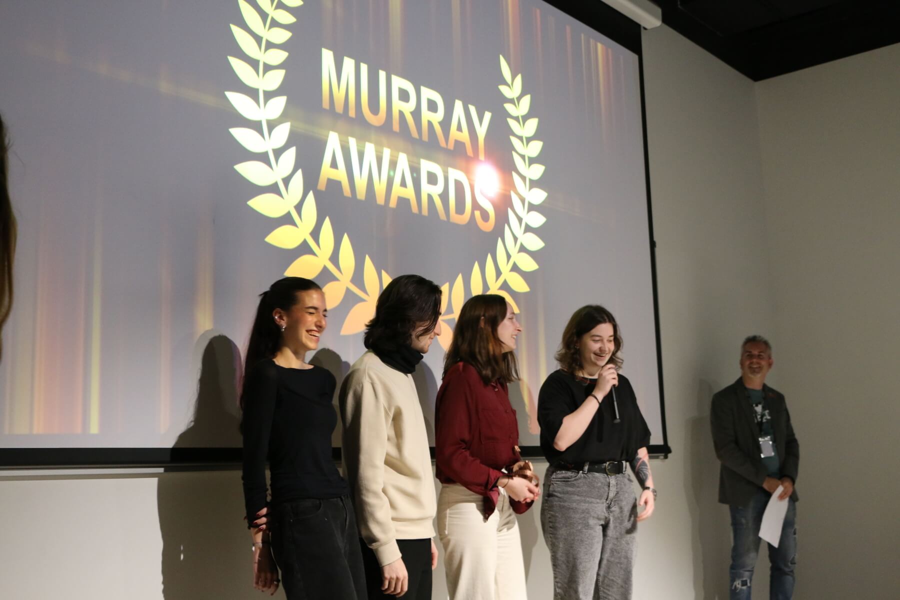 The presenter of the awards show steps aside to allow a student film team a moment to speak after winning an award.